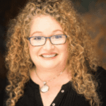 Person wears copper curled hair, glasses, a pendant and black shirt.