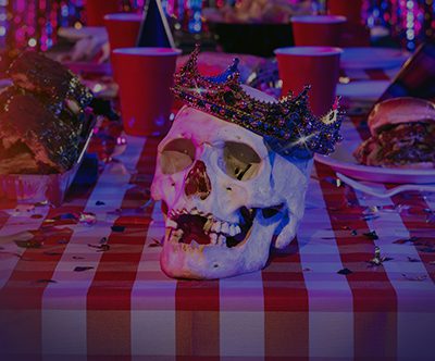 A jewel-encrusted crown adorns a skull sitting on a table littered with red Solo cups and barbecued meats.