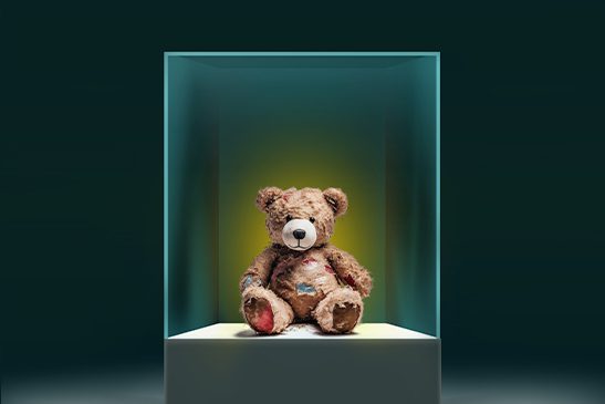 A teddy bear, its fur patchy from years of affection, sits inside a brightly lit glass vitrine as if on display at a museum. 