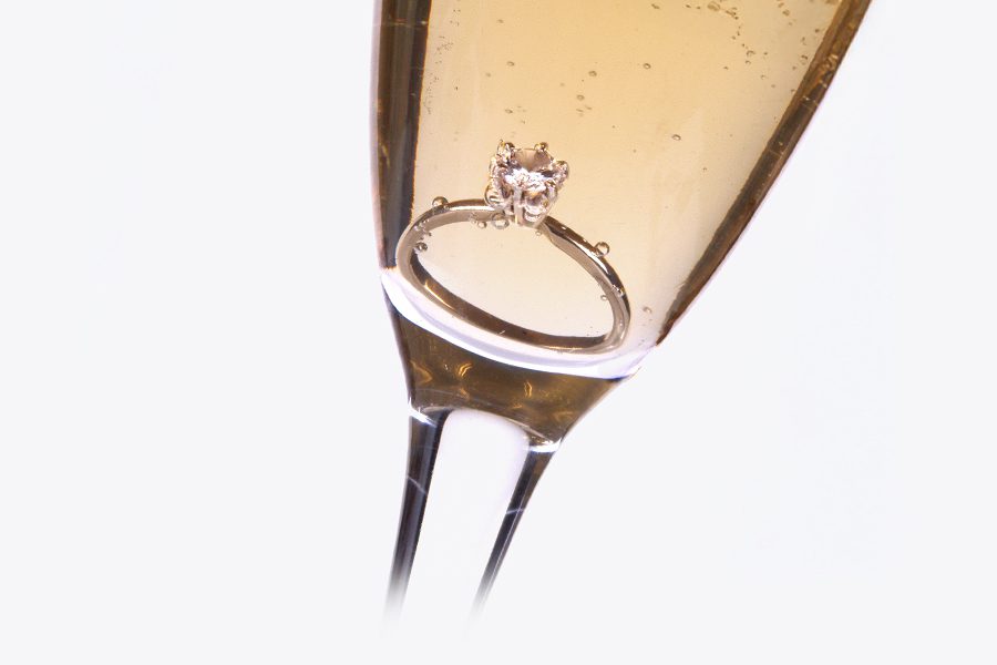 A close-up of a champaign flute filled with amber liquid. At the bottom of the flute sits a diamond on a silver ring.